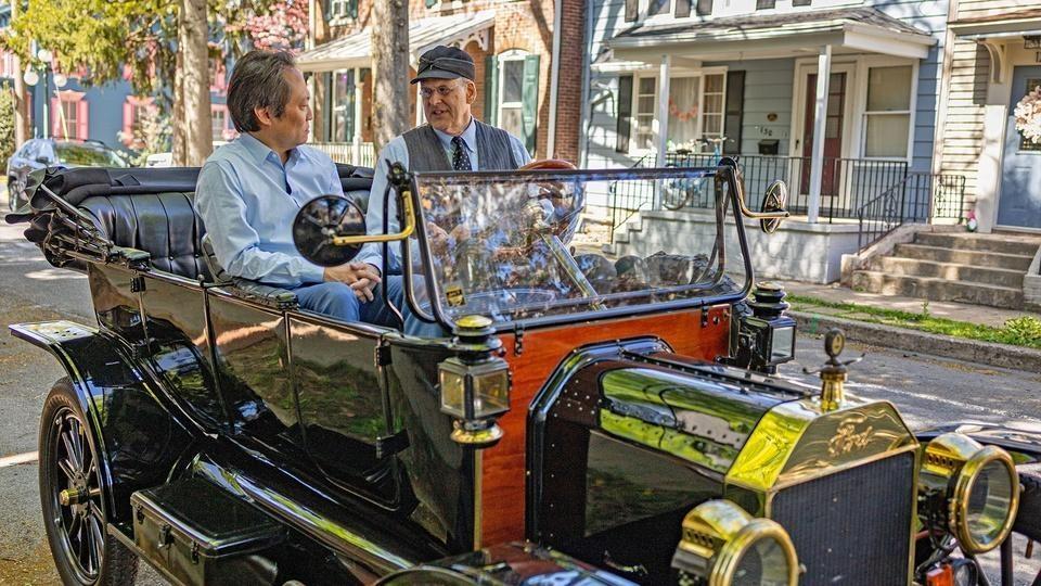 Two men riding in a turn-of-the-century car.
