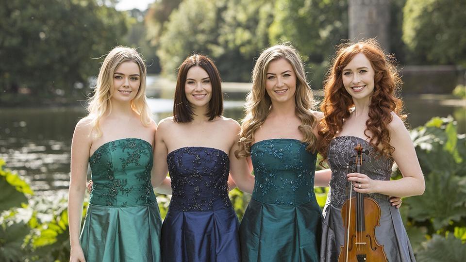 The members of Celtic Woman stand on the edge of a river with a forest and a castle in the background.