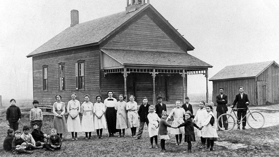 A class of children pose in front of a one-room schoolhouse.