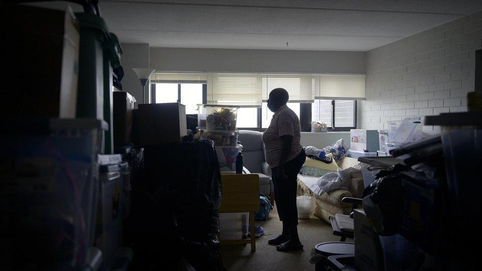 A Black woman stands in a dark room filled with boxes and storage totes.