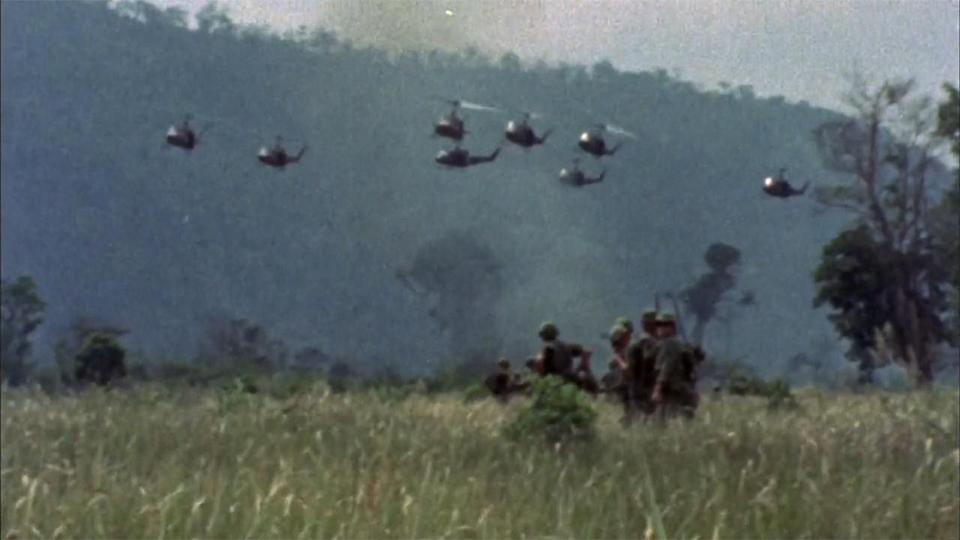 American soldiers walk through a field in Vietnam as helicopters fly across the sky in the distance.