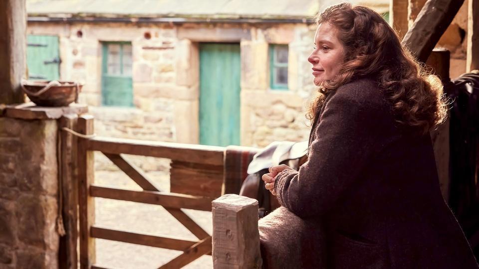 Helen leans on a fence, staring at something out of view.