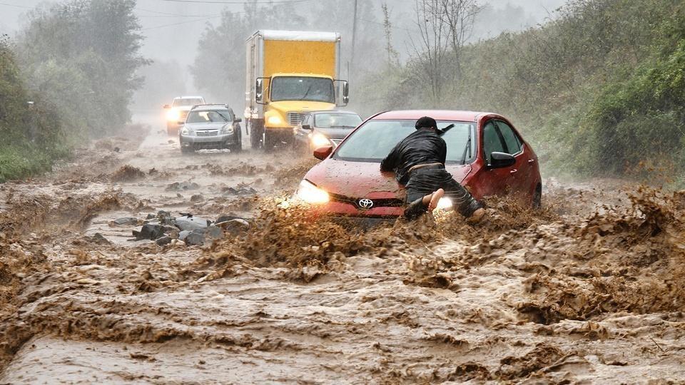 A man clutches to the hood of a car as floodwaters sweep over a road. A semi truck and several other cars stand still further down the road, out of the path of the rushing water.