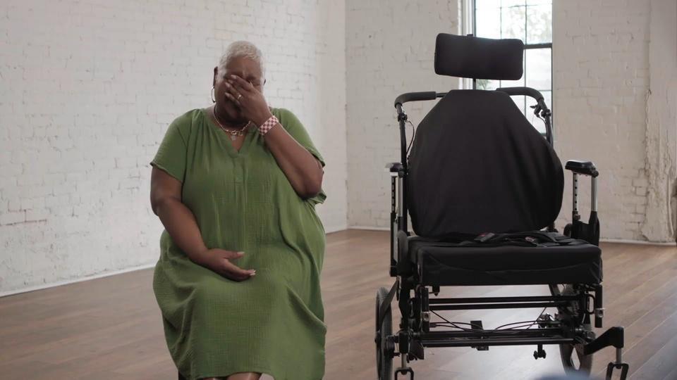 A woman sitting next to a wheelchair cries into her hand.