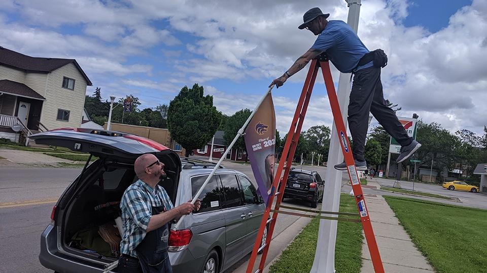 Two men hang a banner with a Bay City Central senior on it from a lamp post.