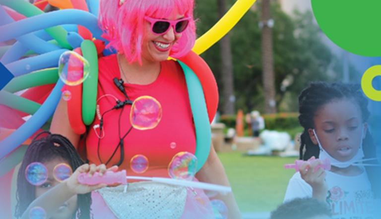 Woman in a pink wig wearing a vest of colorful, elongated balloons watches children blow bubbles.