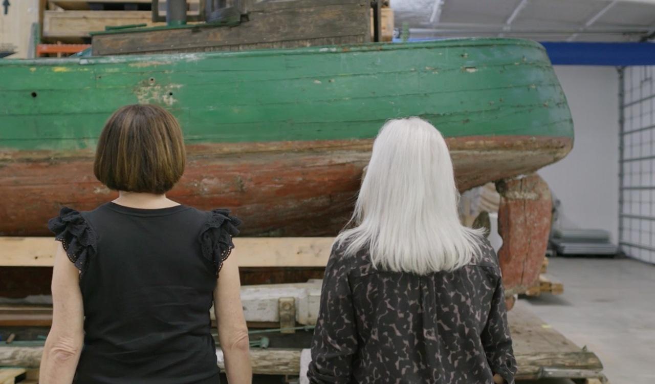 Two women looking at a green boat, Thor. You have a vantage point from behind them.