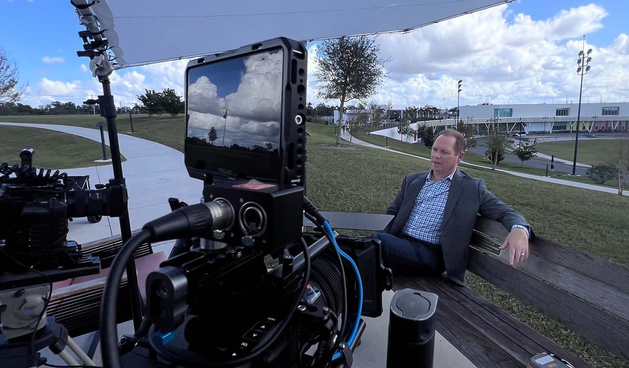 Man sitting on a park bench being interviewed, camera is in the forefront