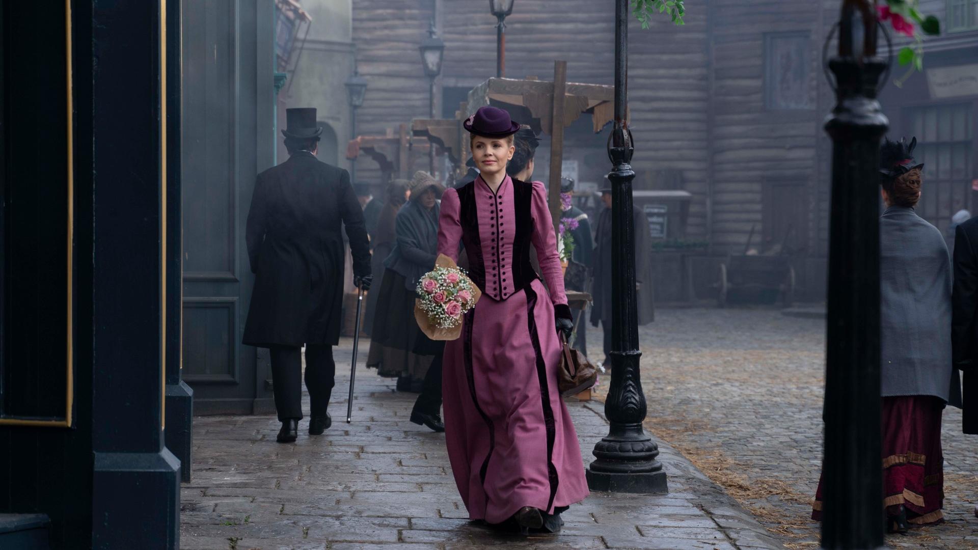 Miss Scarlet walking down a cobbelstone stree well dress with pink flowers, others are around, but there is drama afoot