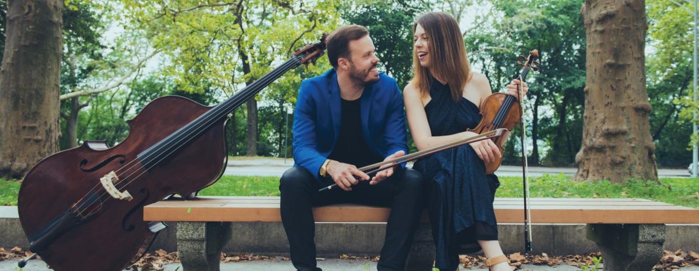 Violinist Tessa Lark and Composer/Bassist Michael Thurber sit on a park bench with their instruments