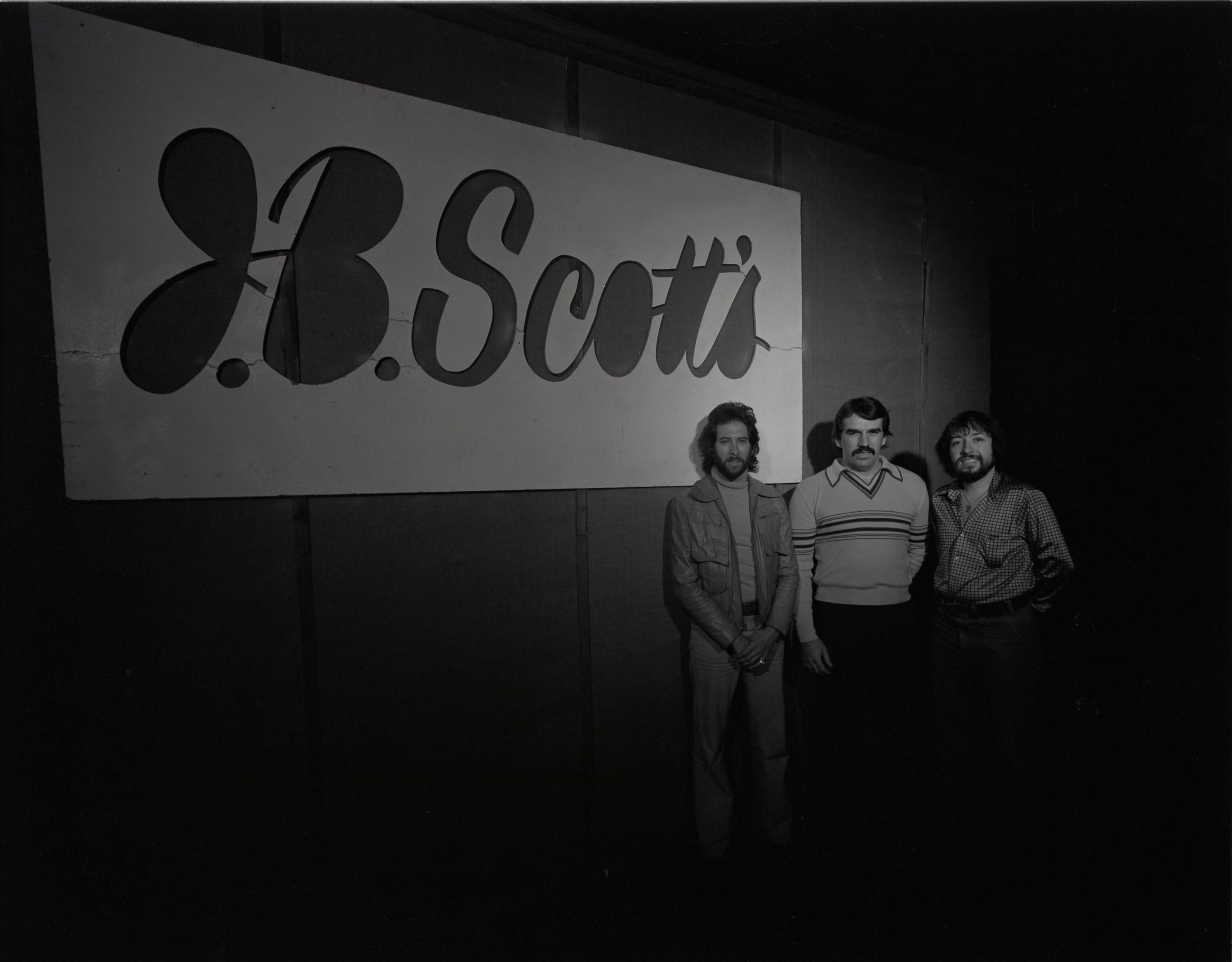 a black and white photo in which three men are standing in front of the J.B. Scott's sign.