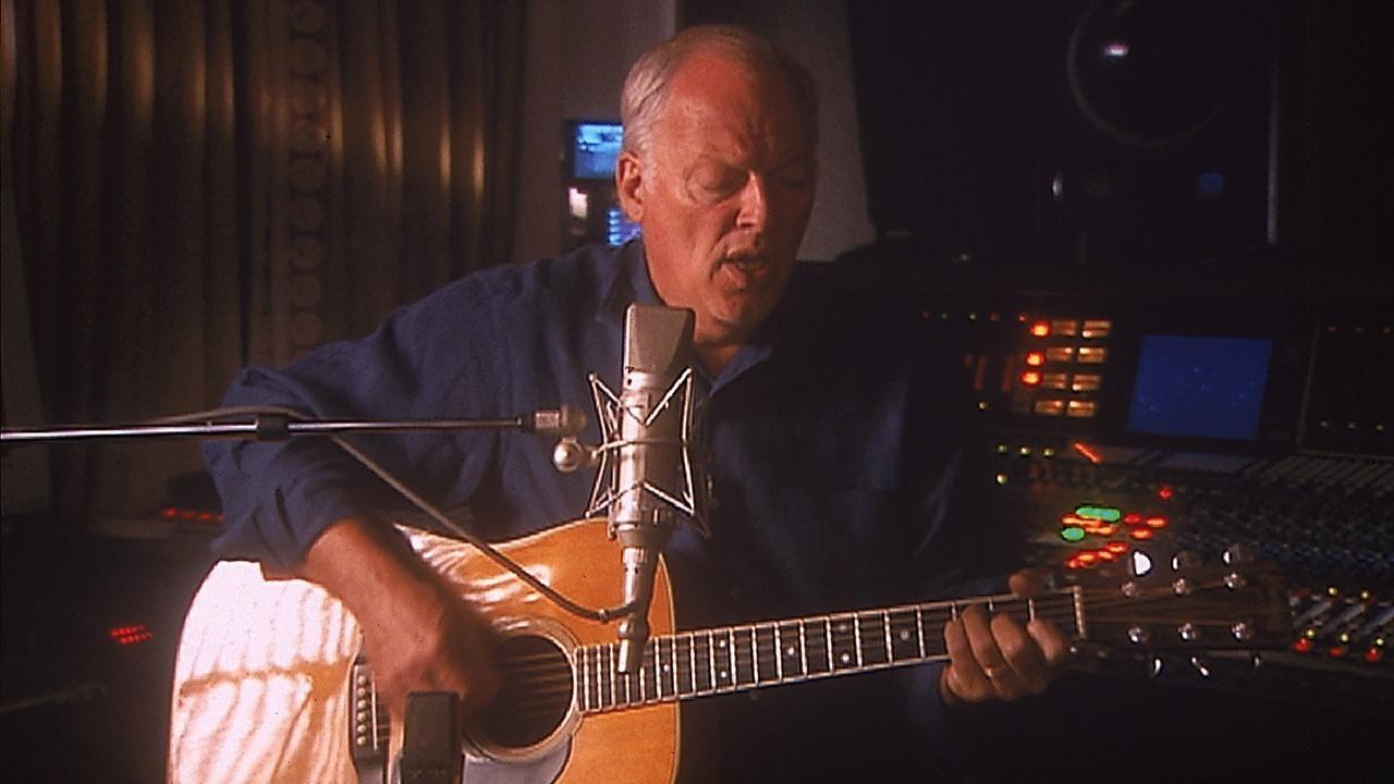 Pink Floyd band member, David Gilmour, sits and plays acoustic guitar while singing into a microphone.