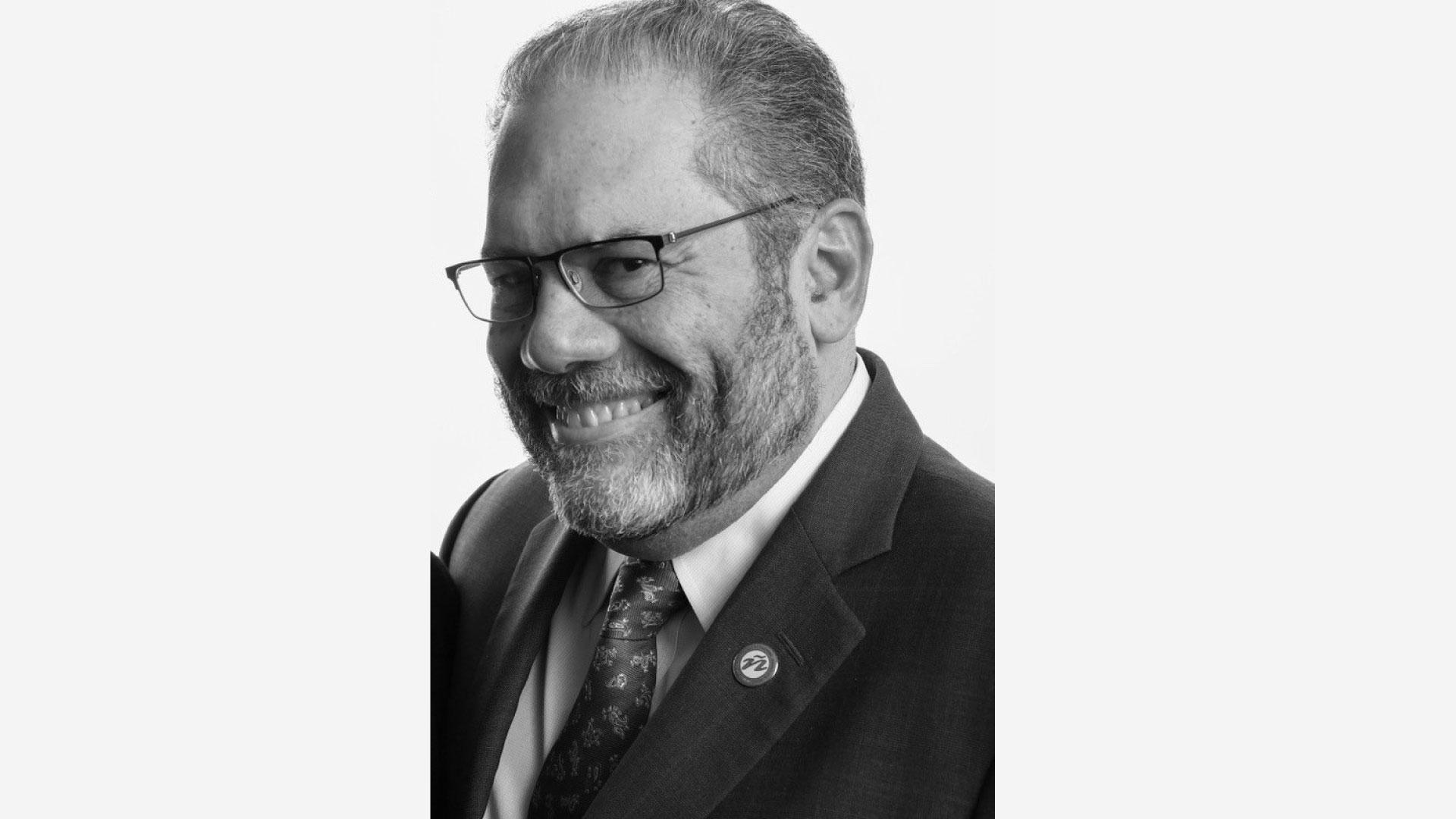 A black and white head shot of Ray Suarez. Ray has a beard and has on glasses while wearing a suit with a collared shirt and necktie.