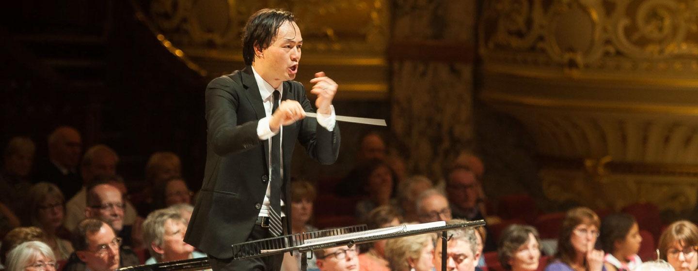 Christopher Tin, wearing a black suit with a white dress shirt, Conducts the Mowbray Orchestra in front of an audience.