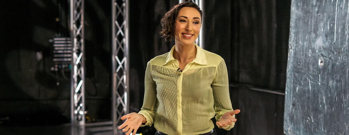 AHA! host Lara Ayad, wearing a yellow blouse, arms held out with hands open, stands angled looking into a camera out of frame.