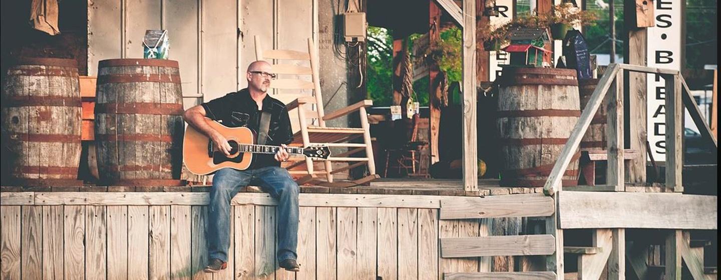 Country Music Singer Billy Montana sits with an acoustic guitar on a wooden porch