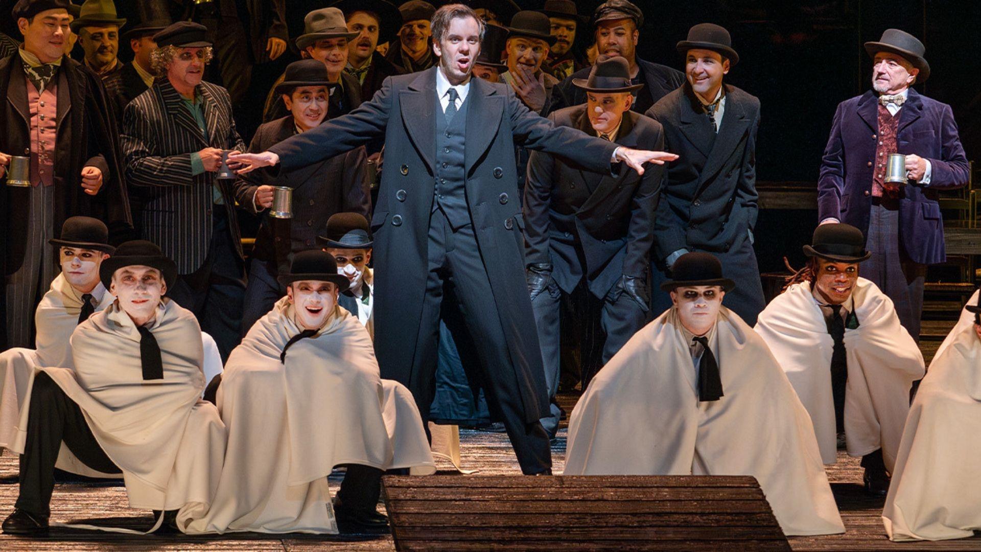 A scene from "Les Contes d'Hoffmann" where a man is singing in a suit, surrounded by tiny men in white capes and black ties