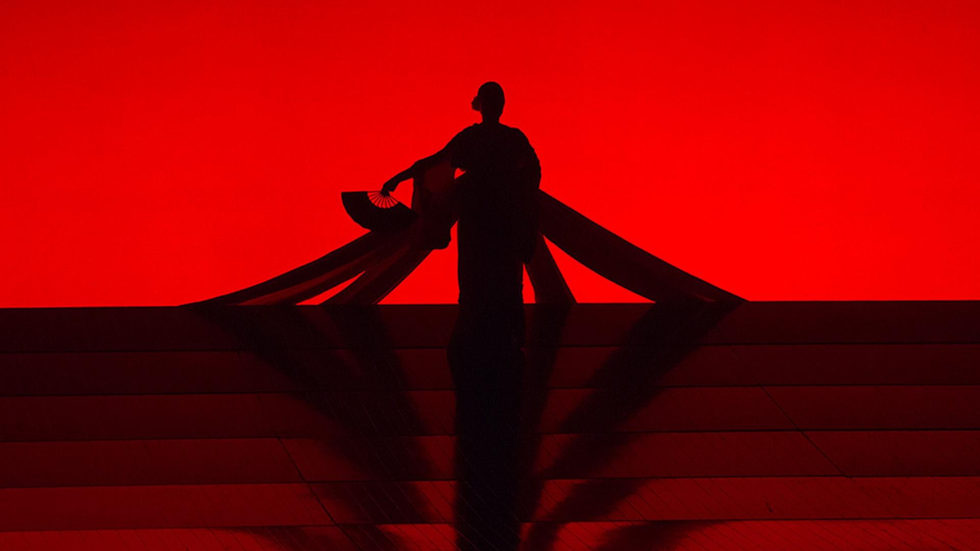 A scene from Madama Butterfly where a shadow of a woman in a kimono holding a Japanese hand fan can be seen with a bright red background