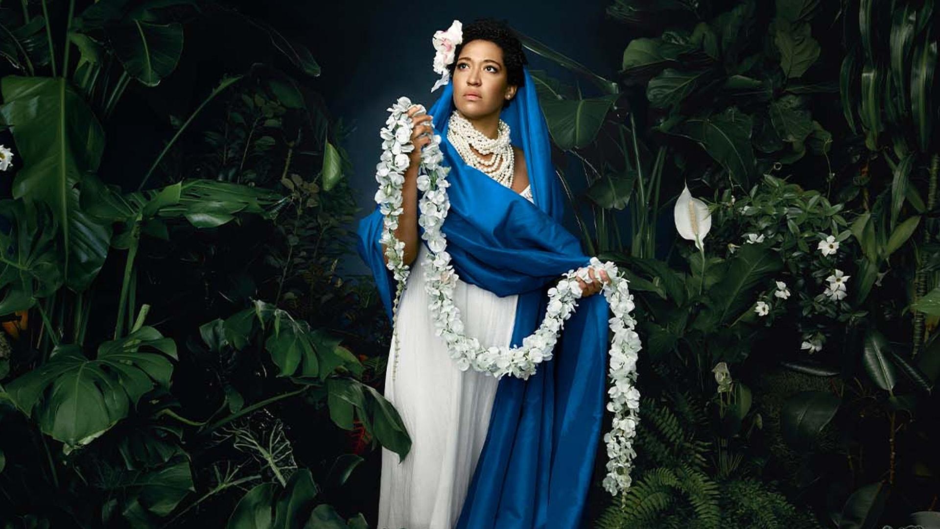 A woman in a white dress with a blue scarf around her and a neck full of pearls clutching a strand of white flowers, with another in her hair