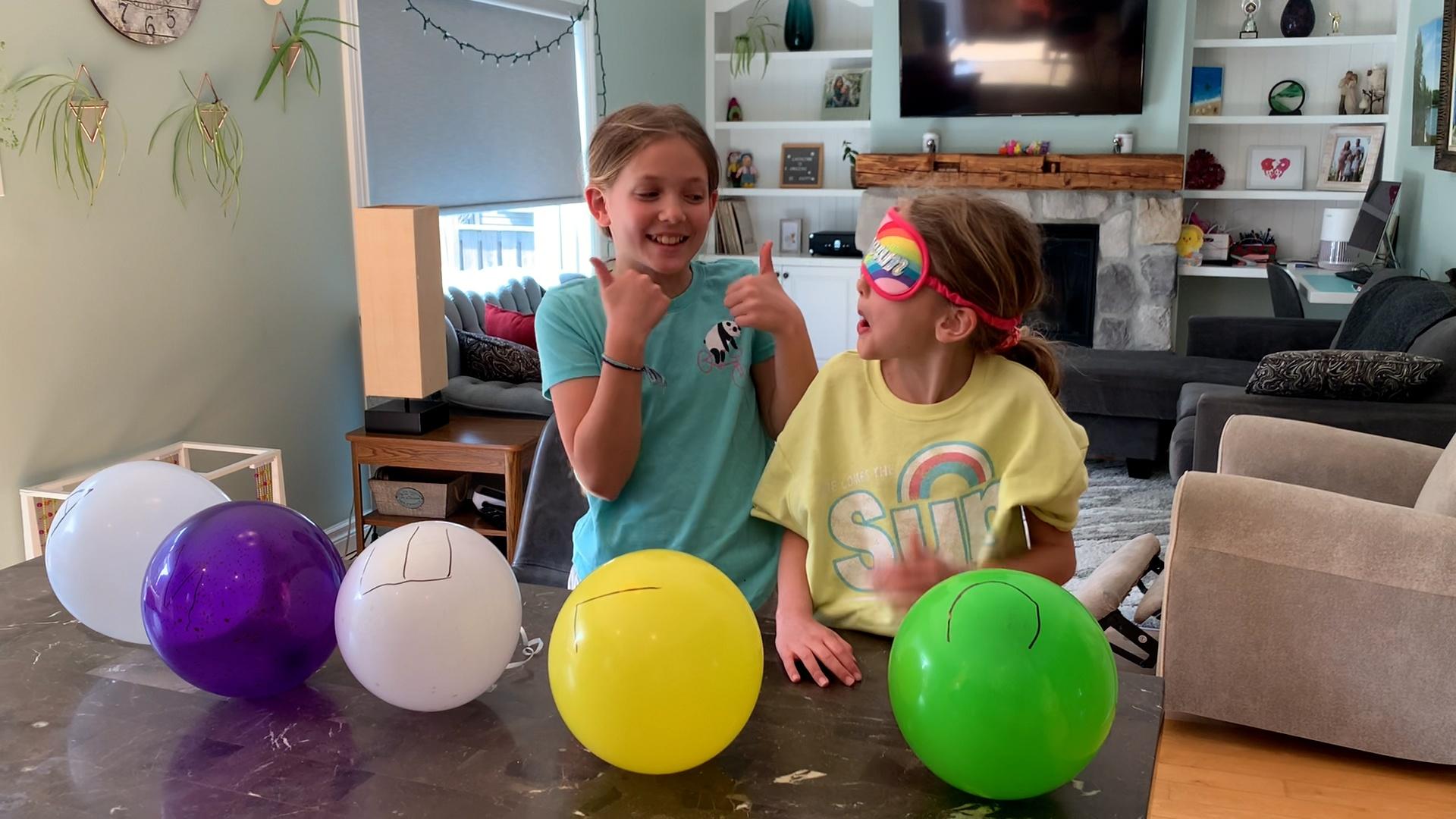 Two young girls, one blindfolded for the challenge and the other smiling with two thumbs up