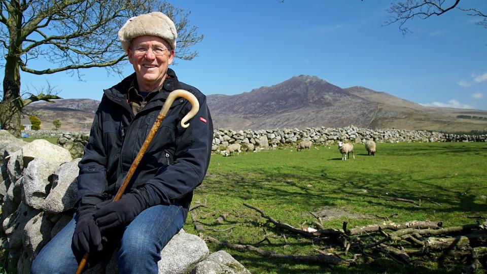 Joseph Rosendo sitting in a rock wall in front of a herd of sheep