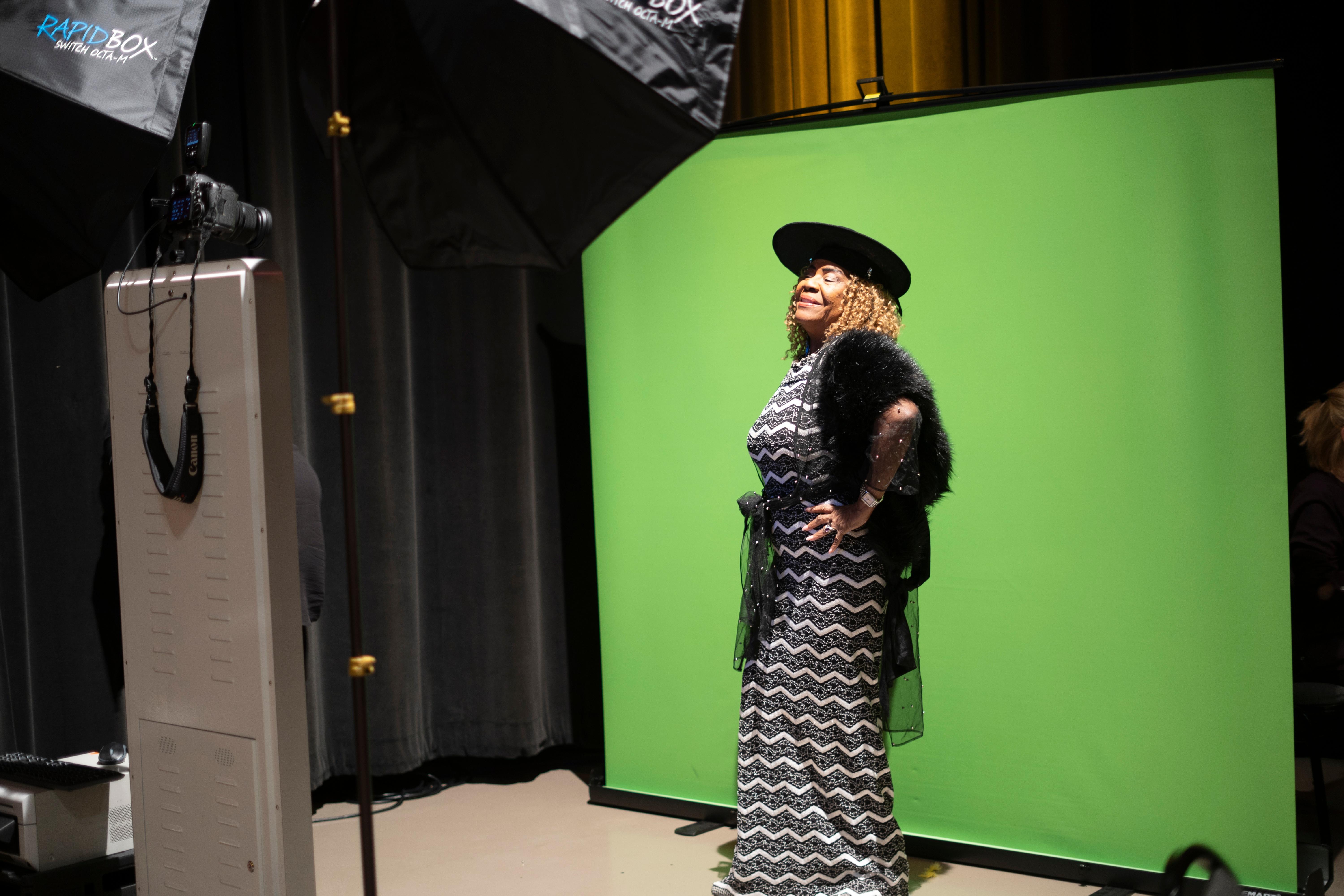 A woman taking advantage of the free photobooth offered at the Senior prom, wearing a black hat, black and white dress, and standing in front of a green screen