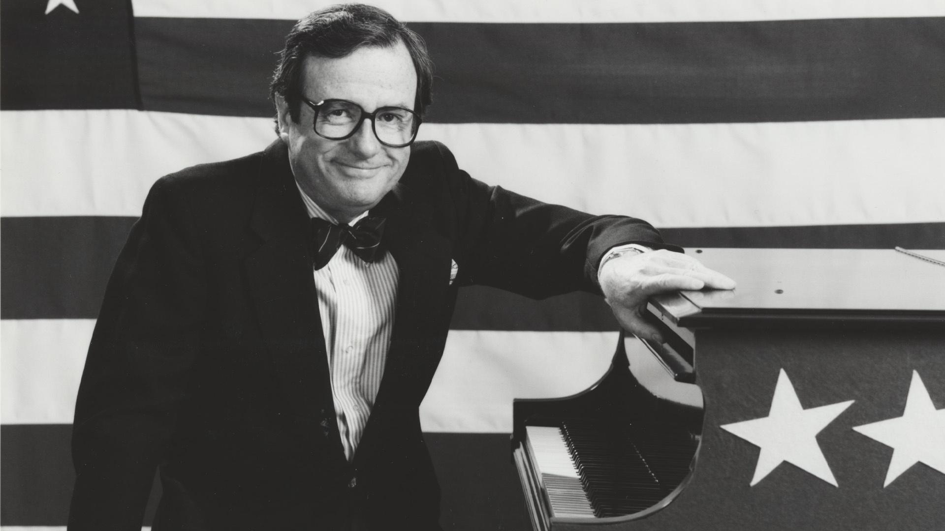 Mark Russell posing at piano with an American flag behind him