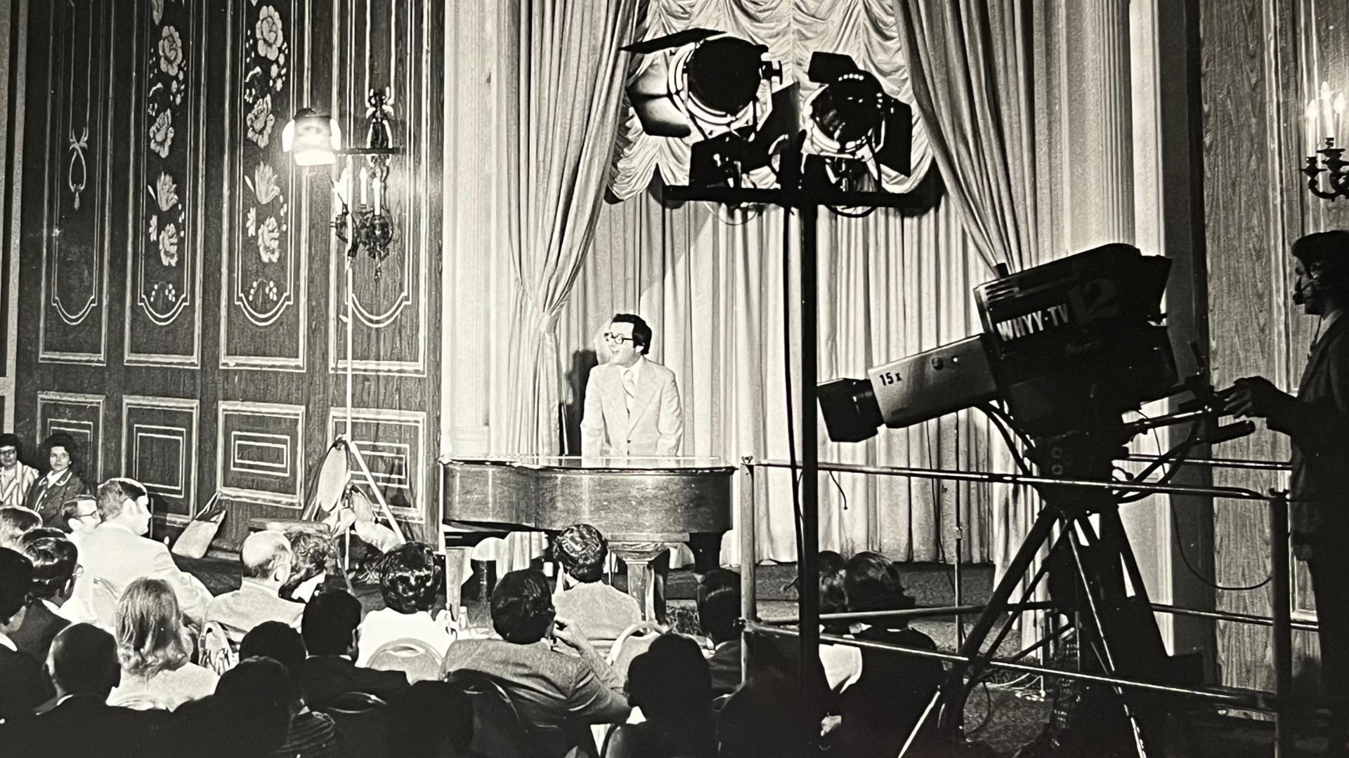 A television camera pointed at Mark Russell performing on stage 