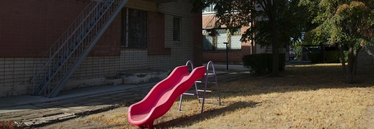 A red slide on dead grass, the surrounding area and buildings are run down