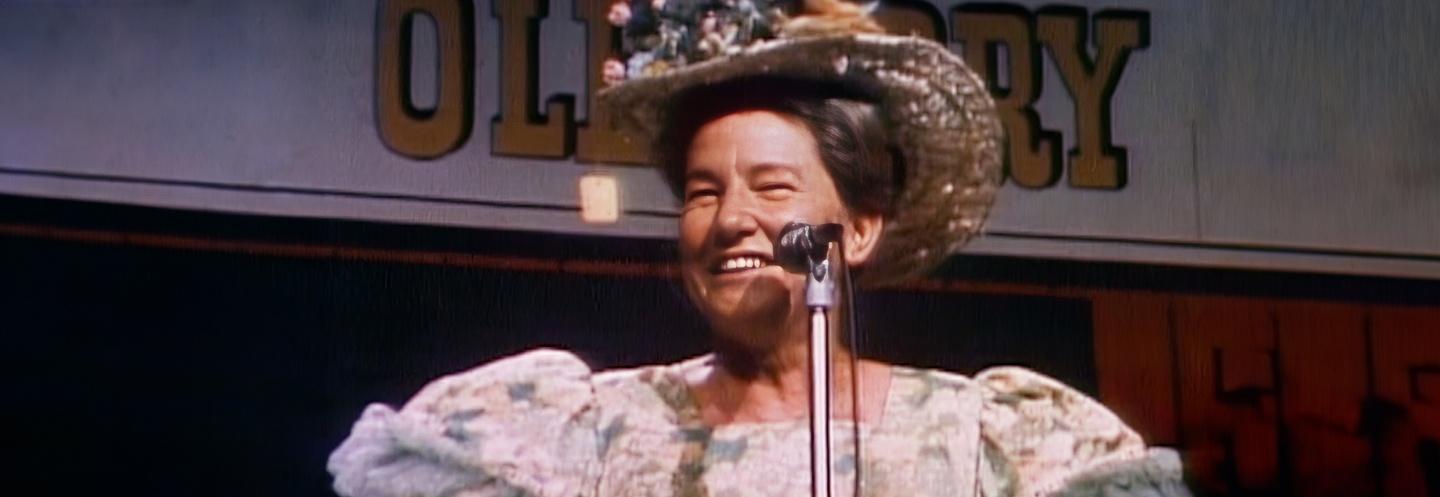 Minnie Pearl wearing a sunhat and puffy sleeved dress, smiling onstage in front of a microphone