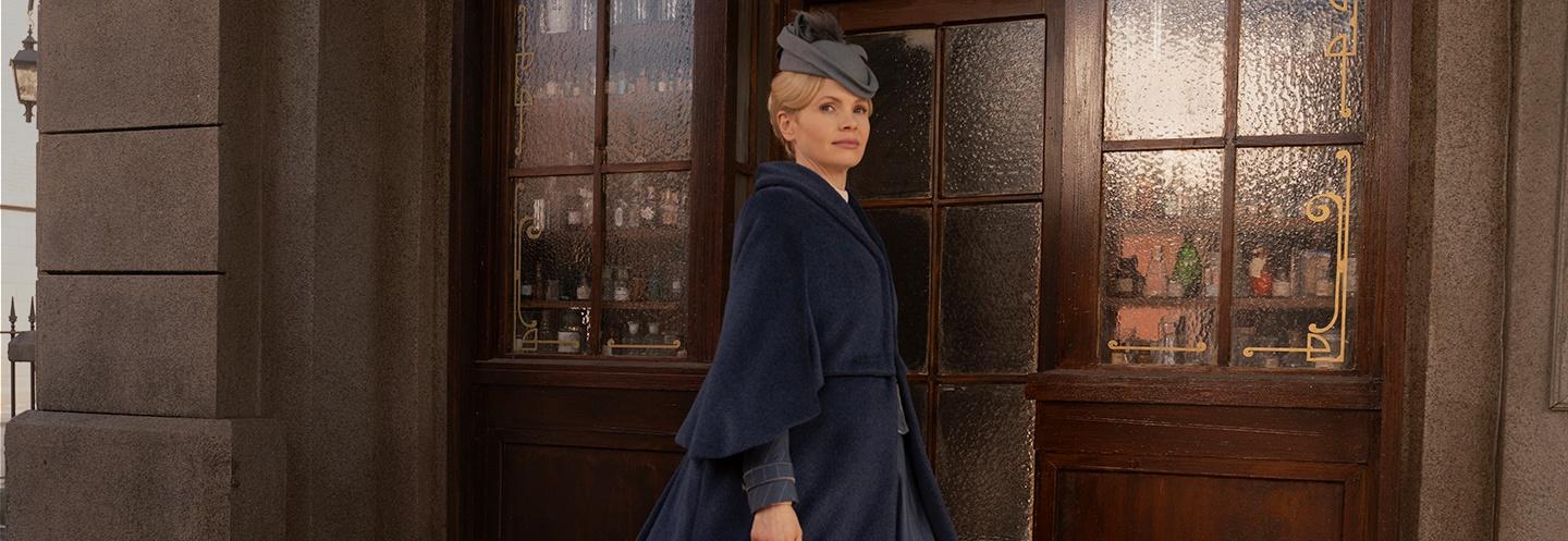 "Miss Scarlet" standing in front of a doorway in a long blue coat and matching hat