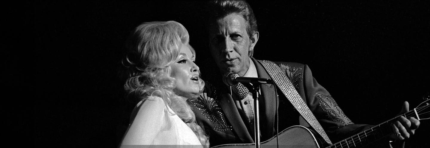 Black and white image of Dolly Parton and a male singer singing into a shared microphone