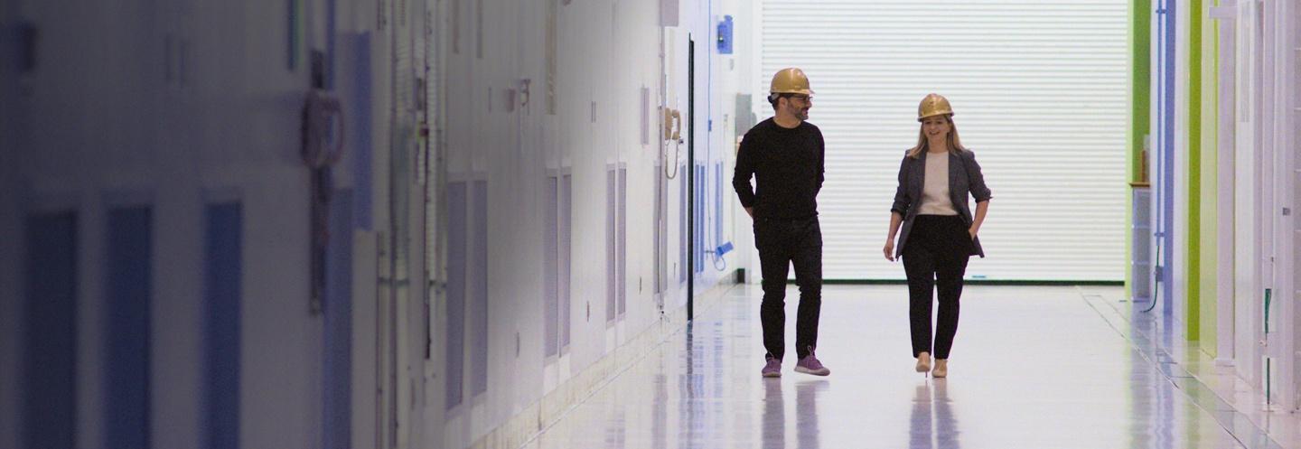 2 people walking down a hallway wearing yellow hard hats