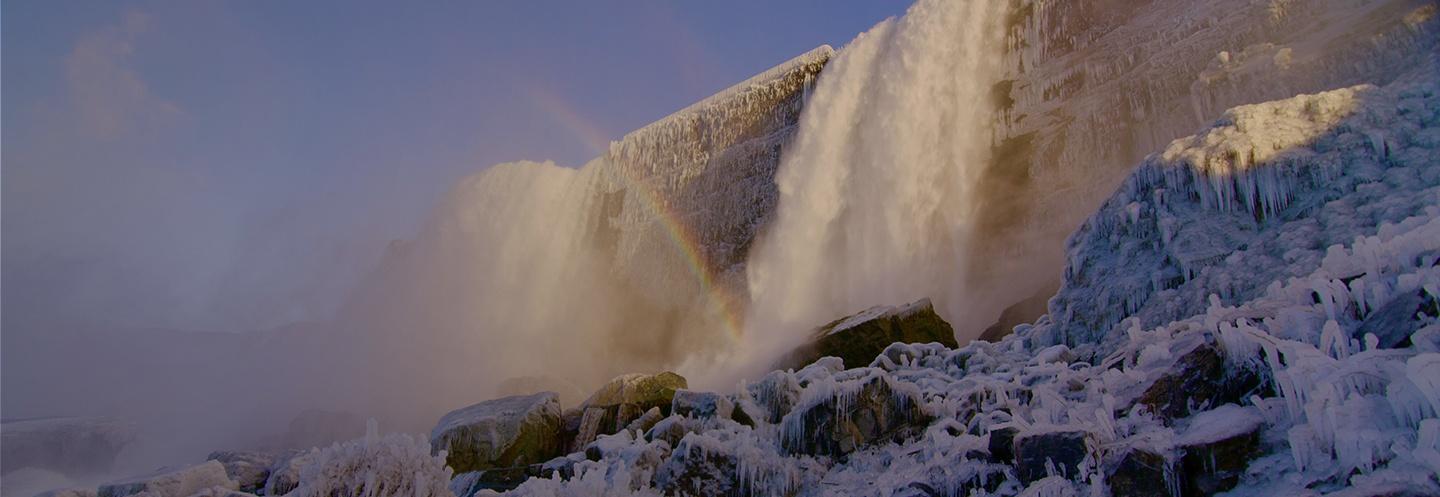 Photo of an icy Niagara Falls take from the bottom, and a rainbow is partially visible