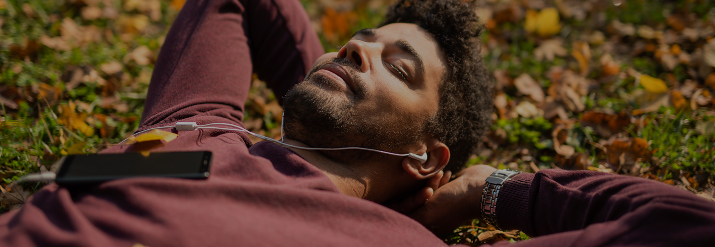 A man laying down in grass with fall leaves. His arms are up behind his head, his eyes are closed, and he is wearing wired ear buds