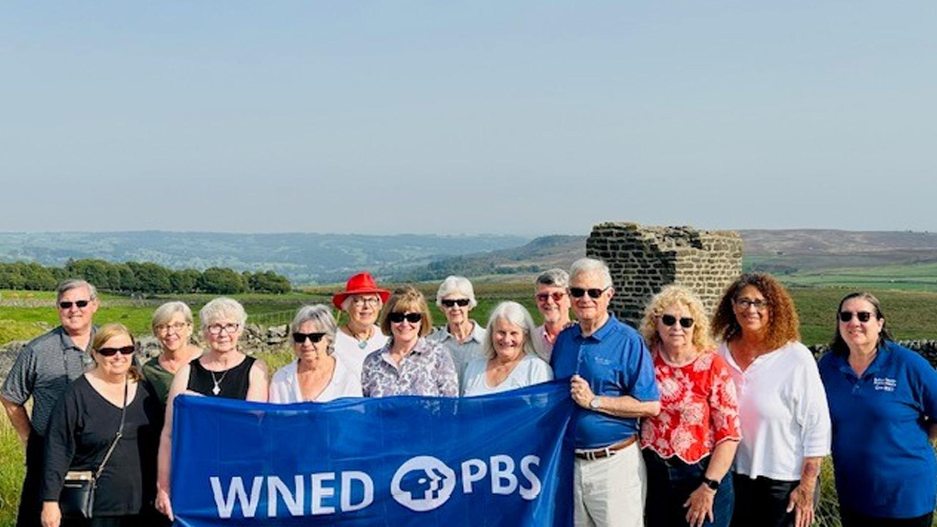 group holding WNED PBS flag with the Yorkshire Dales in the background while on the All Creatures Great an Small tour of England