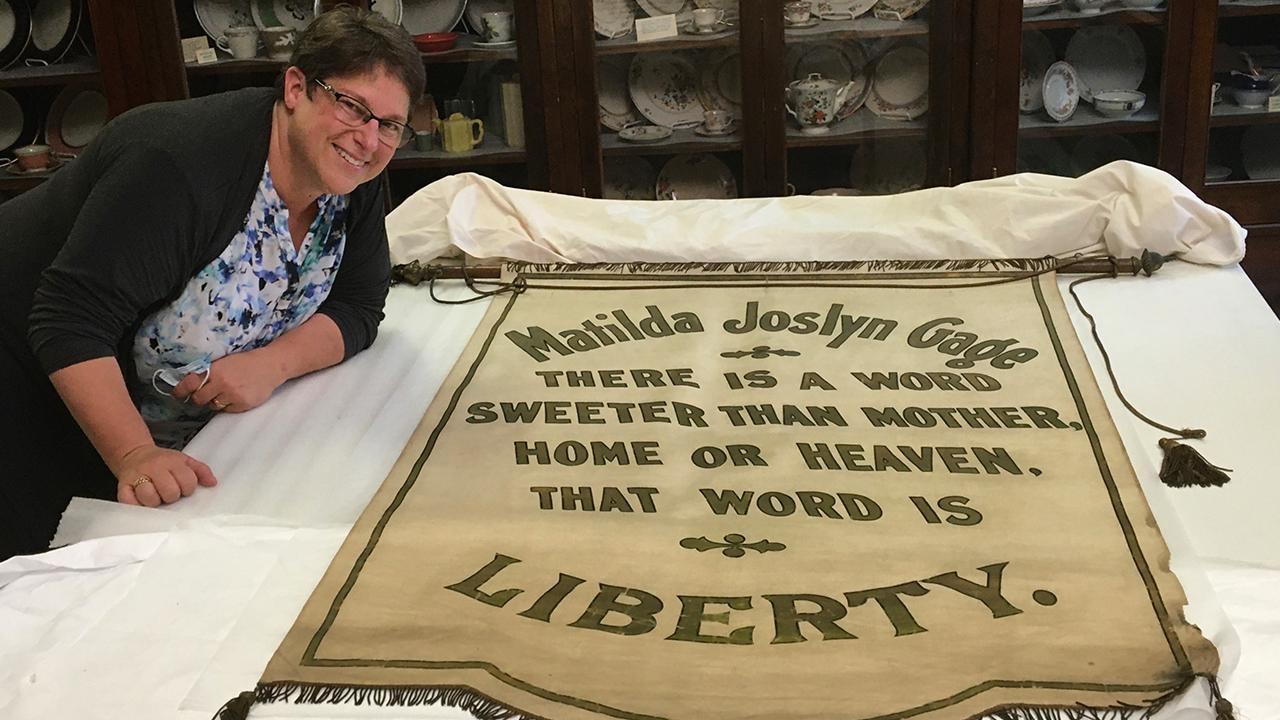 Lynne Bader poses in front of a Matilda Joslyn Gage banner