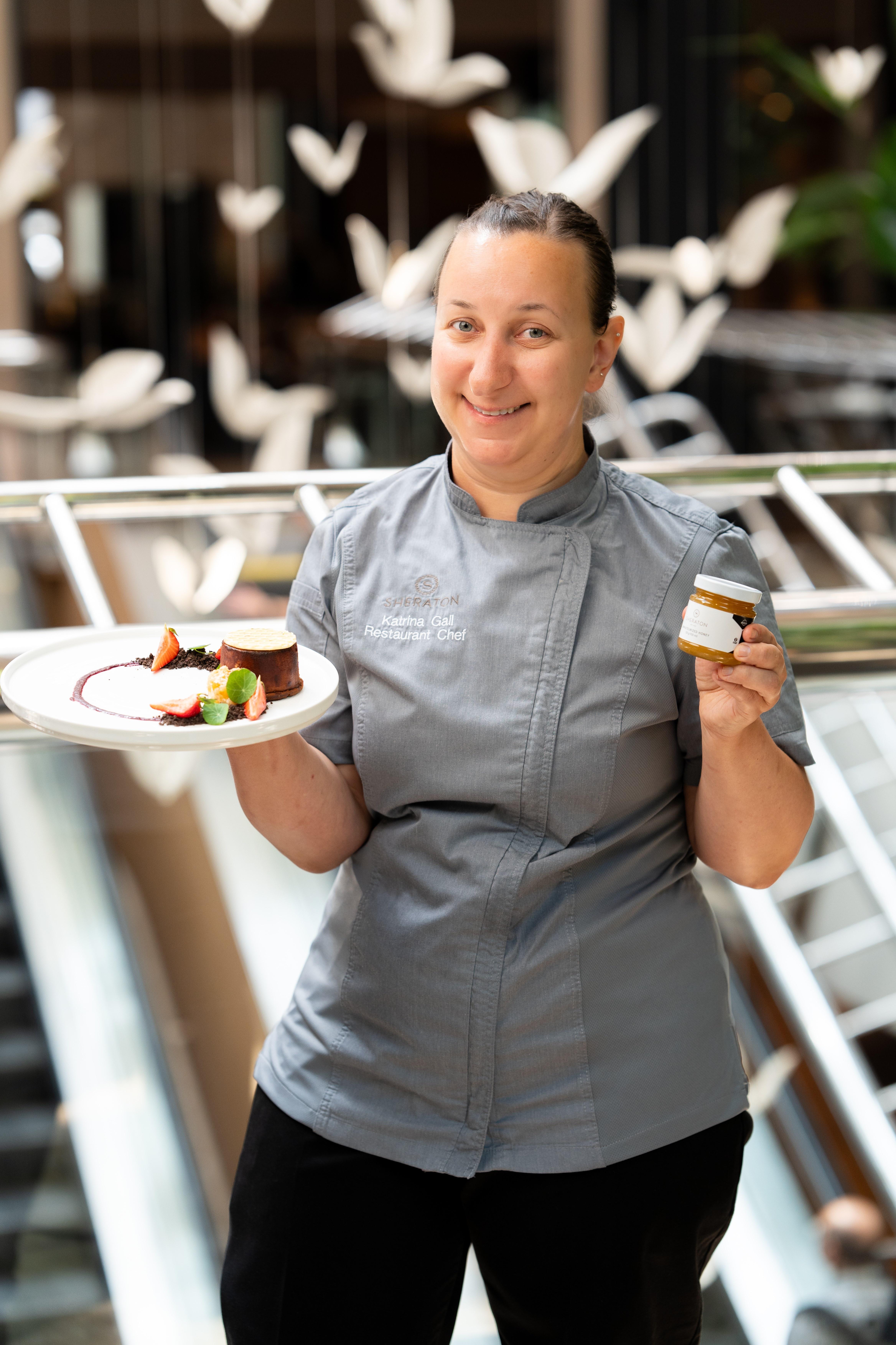 Chef Katrina Gall standing and smiling with a plate of food and a small jar.