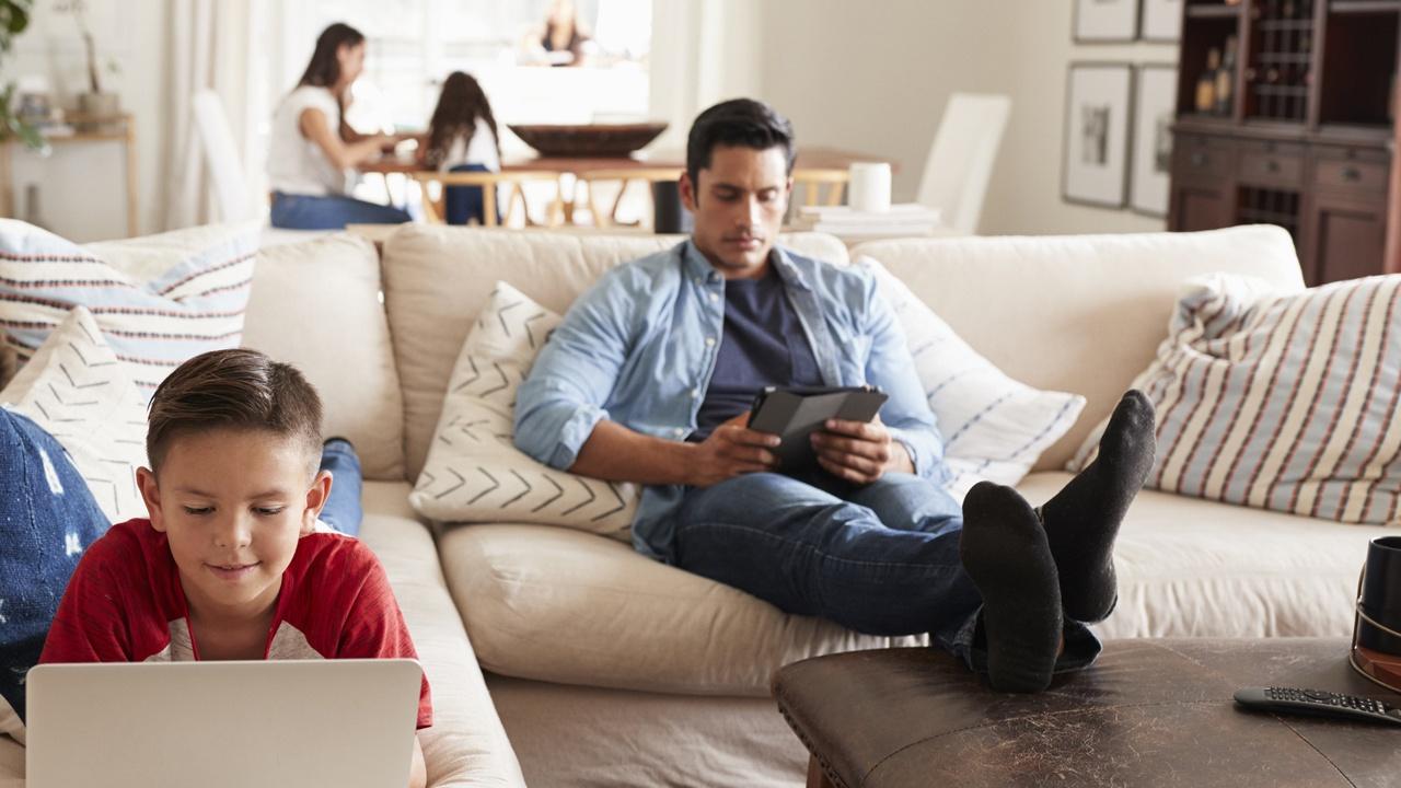 father on ipad and son on laptop in living room , while mom and daughter sit at table in background