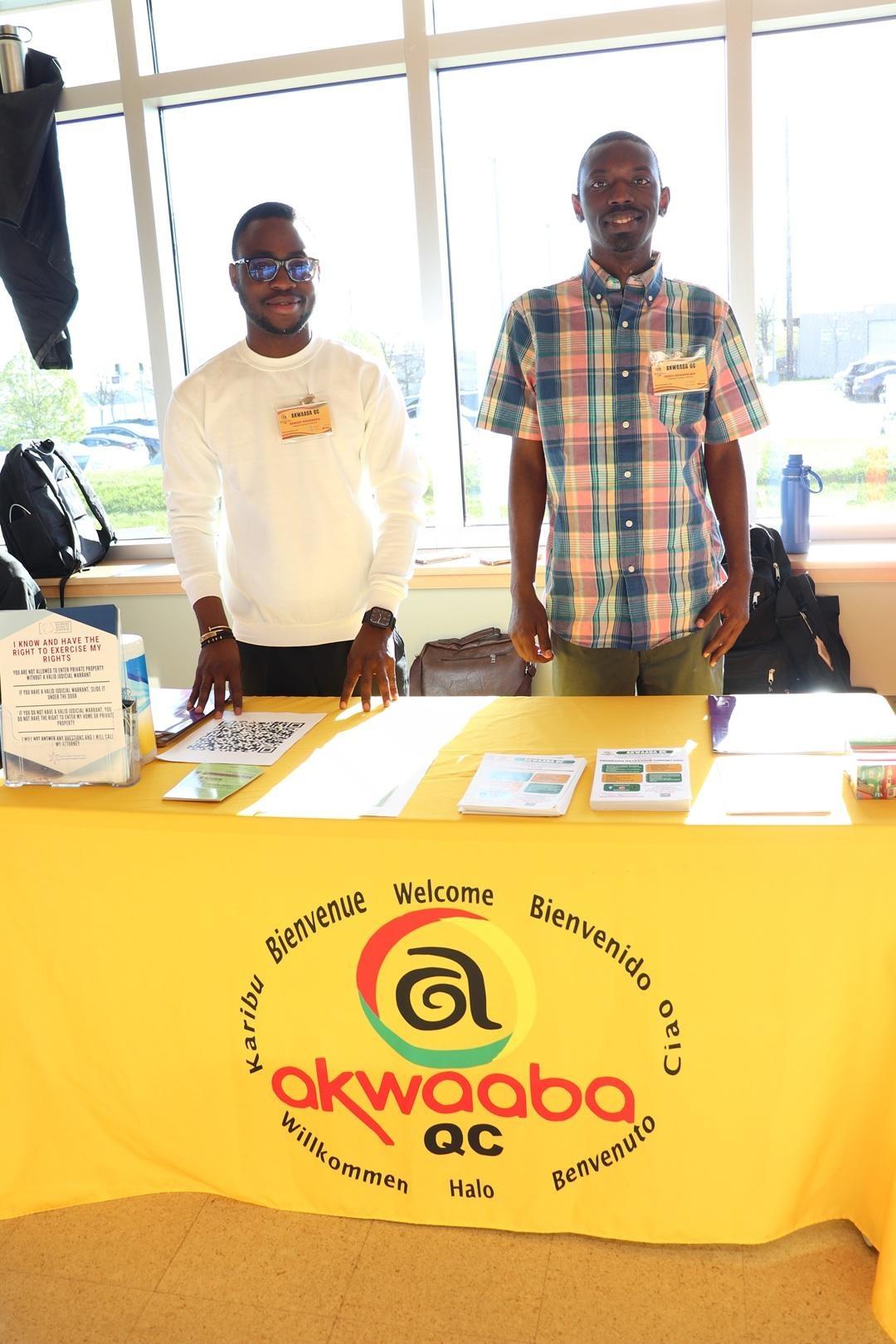 Two men stand in front of large windows manning a table dressed in a yellow cloth branded with the Akwaaba QC logo and messages of "Welcome" in multiple languages