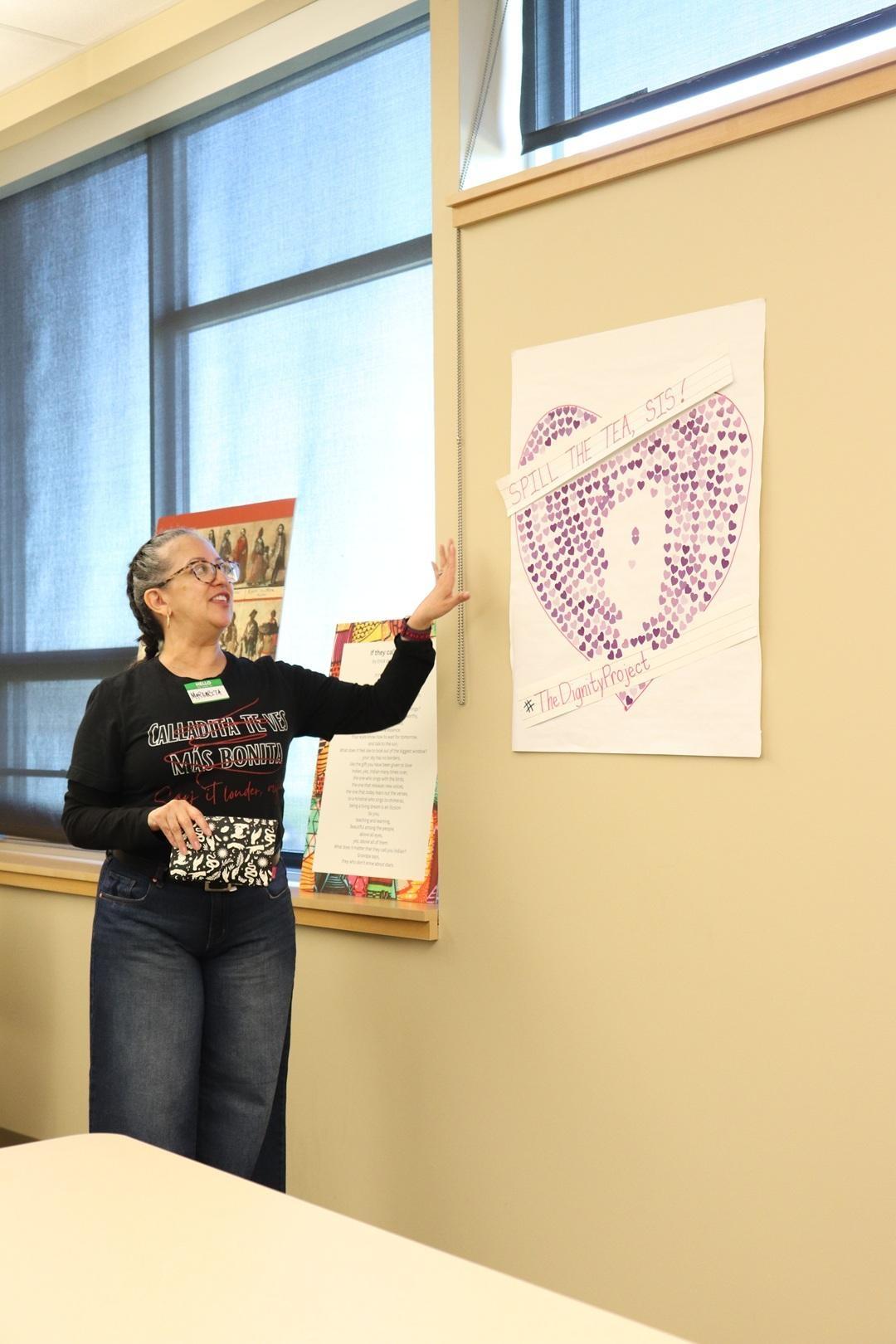 A teacher wearing jeans, a dark long-sleeved shirt, and glasses stands in front of a window pointing at a drawing affixed to a beige wall