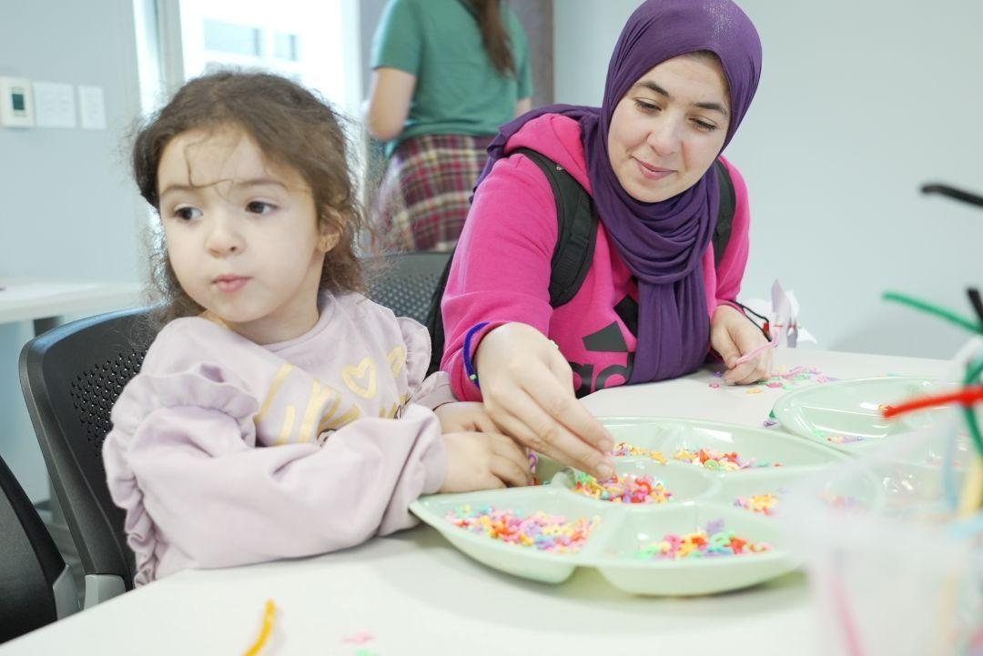 A woman wearing a pink shirt and a purple hijab sits at a table helping a child with small craft items in a round tray