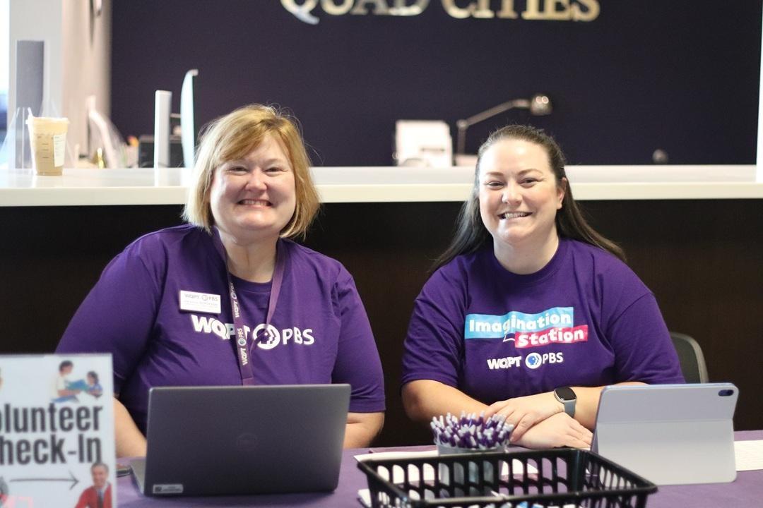Two volunteers sitting at a table wearing purple Imagination Station WQPT PBS t-shirts