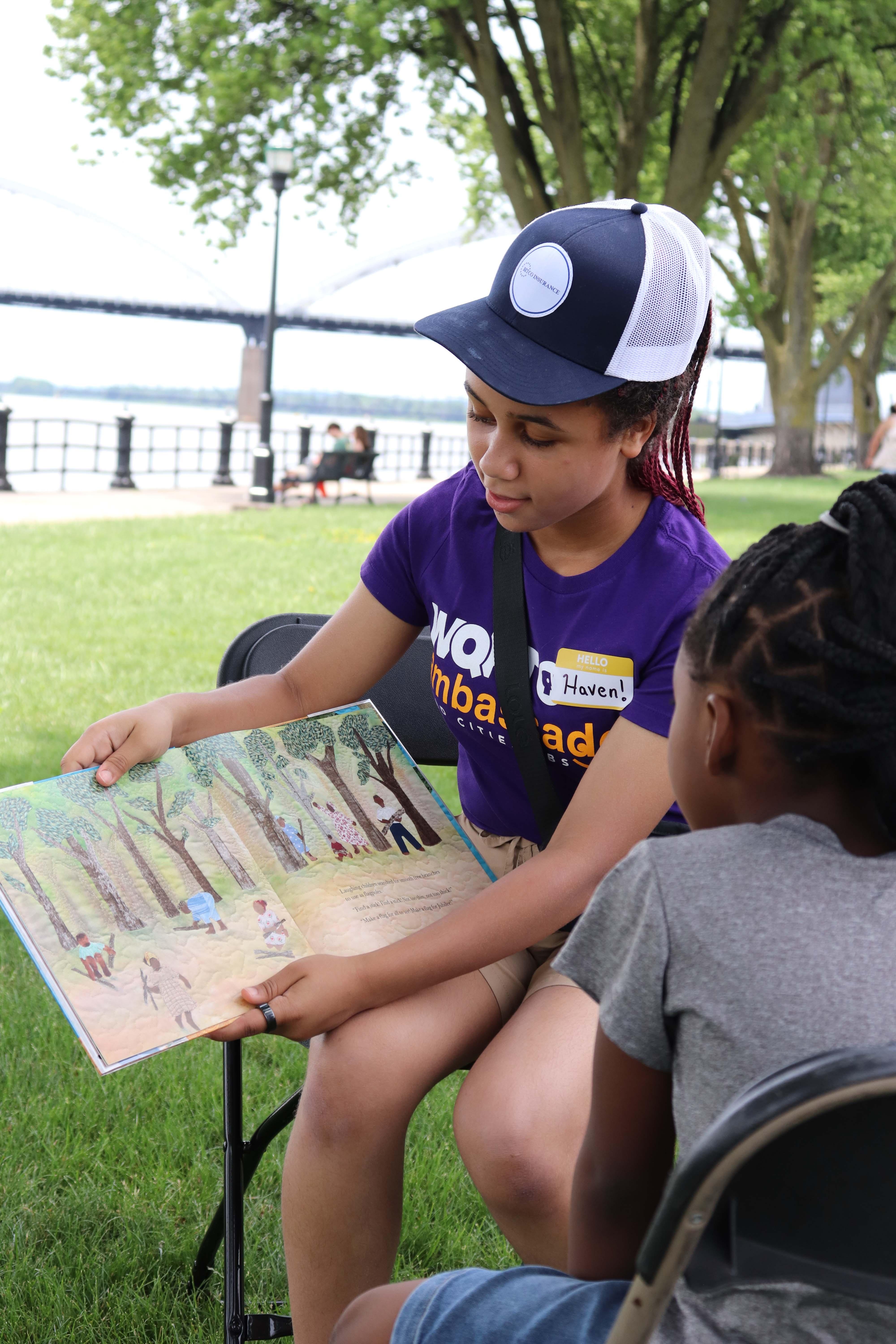 A WQPT Ambassador wearing a purple t-shirt and baseball cap holds a picture book while sitting on a lawn with a child