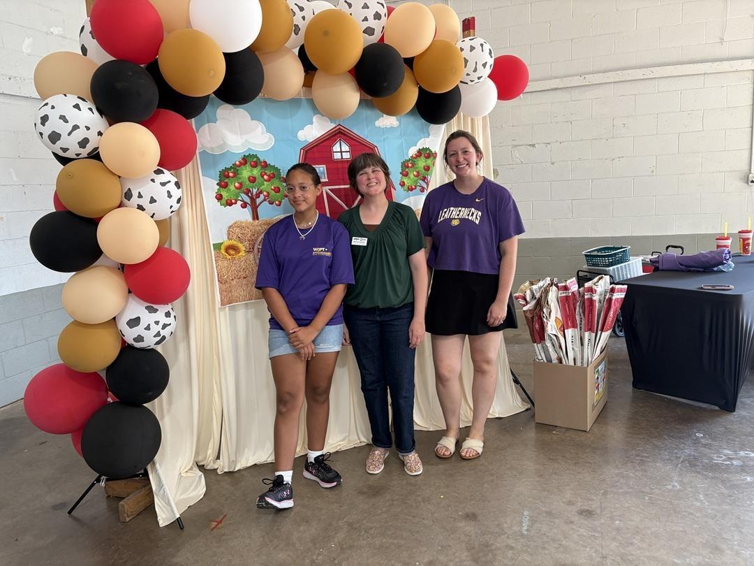 Three people stand posing in front of a backdrop lined with black, red, beige, brown, and white spotted balloons