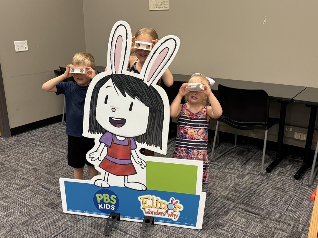 Three children stand behind an "Elinor Wonders Why" cardboard cutout, looking through white card stock viewers