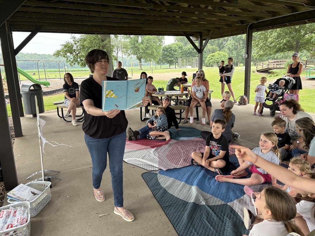 A woman shows an open book to a group of children sitting on a blanket under an outdoor canopy