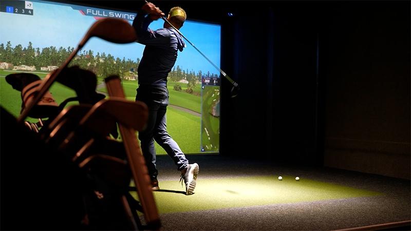 Golfer follows through on a swing in an indoor simulator, with golf clubs in the foreground.