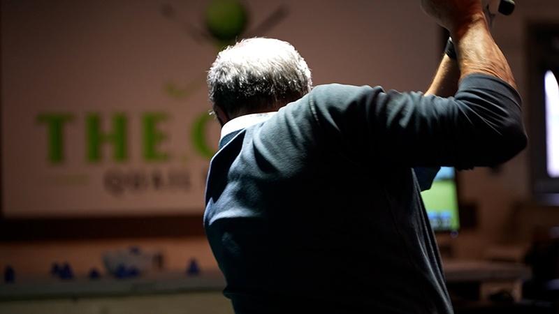 Close-up from behind of a gray-haired golfer with arms raised during an indoor golf swing.