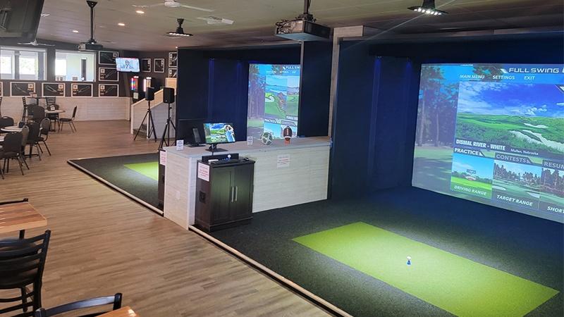 Two indoor golf simulator bays with large projection screens, green hitting mats, and nearby seating.